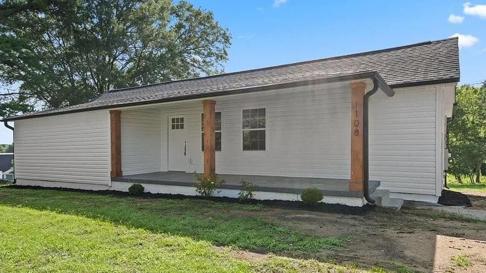 Three-quarter view from the front yard, where the new dark grey architectural shingles run unbroken from the ridge down over the full-width front porch to a fresh drip edge and gutter along the eave.