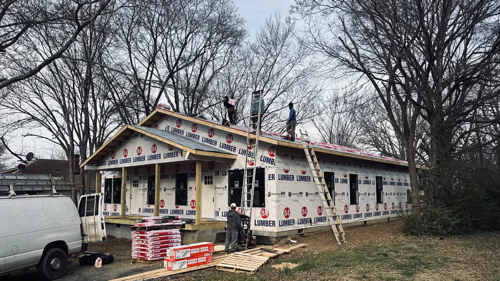 A Rixa crew at work on the roof of a single-story house: the deck is covered in grey synthetic underlayment, wrapped shingle bundles are staged along the ridge, two extension ladders lean against the roof, and more bundles wait on pallets in the yard.